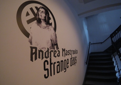 Czarno – biała grafika na ścianie przedstawiająca postać w długich włosach na tle logotypu CSW Łaźnia. Pod postacią widoczny jest napis „Andrea Mastrovito - Strange Days”