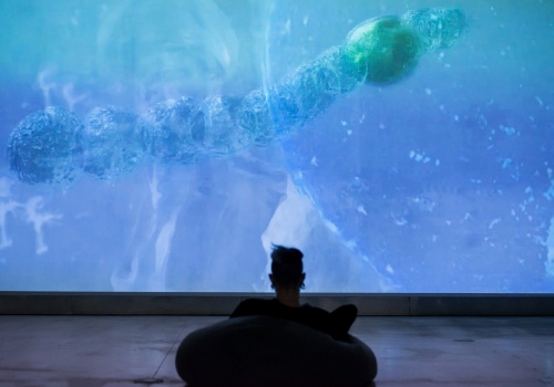 6.	Ciemna sala wystawiennicza. Zbliżenie na ścianę, na której wyświetlana jest wielkoformatowa praca audiowizualna, ukazująca plankton w powiększeniu.