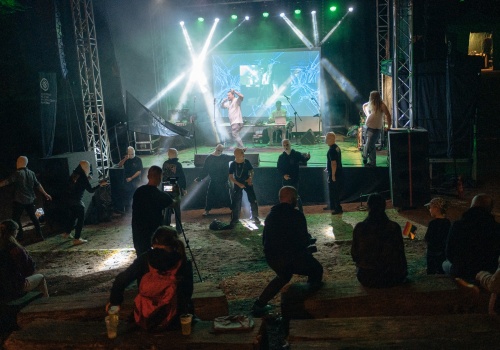 Kadr na oświetloną scenę Teatru Leśnego, w trakcie trwania koncertu zespołu „Korozja”. Pod sceną widoczni są zamaskowani tancerze.