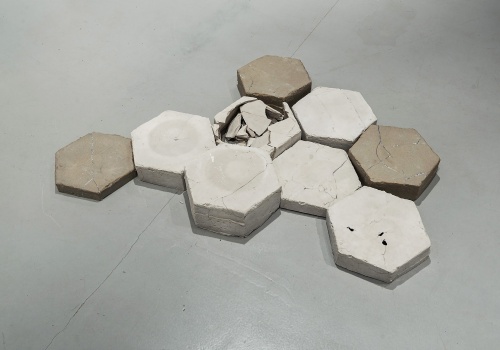 Dziewięć heksagonalnych płyt odlanych z materiału budowlanego, ustawione na podłodze. Jeden z heksagonów jest uszkodzony.