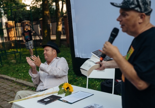 Kadr na Krzysztofa Skibę mówiącego przez mikrofon. W prawej dłoni trzyma otwartą książkę. Obok przy stole siedzi Paweł Konjo Konnak.