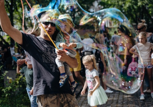 Zdjęcie, mężczyzna z dzieckiem na rękach puszcza ogromne bańki przy pomocy sznurka do tworzenia baniek mydlanych. Za nimi znajduje się liczna grupa osób stojąca w kolejce do festiwalowych atrakcji.