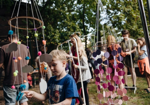 Zdjęcie, licznie zebrani festiwalowicze wykonują kolorowe karuzele z włóczki i papieru. W kadrze znajdują się dwie gotowe prace. Karuzelą po prawej stronie kadru bawi się chłopiec.