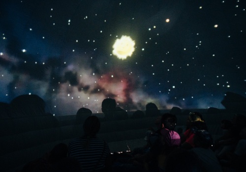 Zdjęcie, wnętrze ciemnego namiotu, w którym wyświetlany jest film Mobilnego Planetarium Cassiopeia. Dzieci siedzą na podłodze. Nad ich głowami znajduje się rozświetlone gwiazdami niebo.