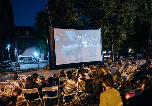Zdjęcie, liczna grupa festiwalowiczów siedząca na leżakach, kocach i pufach ogląda film na podwórku CSW ŁAŹNIA 2 w Nowym Porcie. Film wyświetlany jest na dużym, mobilnym ekranie.