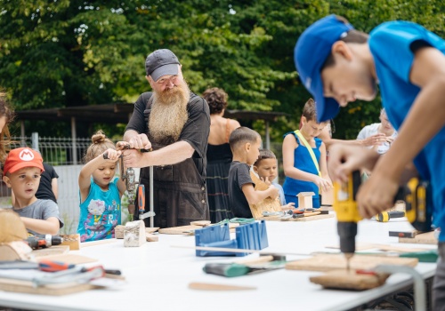 Zdjęcie, w kadrze znajduje się fragment stołu warsztatowego wraz z uczestnikami festiwalu. Dzieci wykonują pirackie statki z drewna. W centrum kadru znajduje się mężczyzna z długą, rudą brodą, ubrany w czarny, roboczy kombinezon, który pomaga dziewczynce konstruować zabawkę.