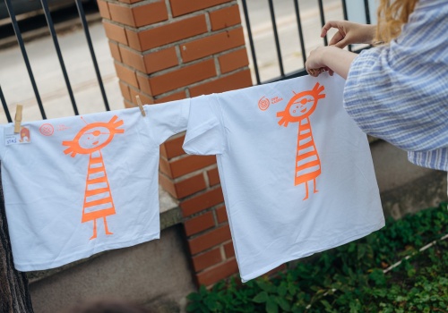 Zdjęcie, zbliżenie na białe koszulki rozwieszone na lince. Na koszulkach znajduje się pomarańczowa grafika z Łaźniakiem, która schnie. W kadr po prawej stronie wkrada się sylwetka dziewczyny, która rozwiesza jedną z koszulek.