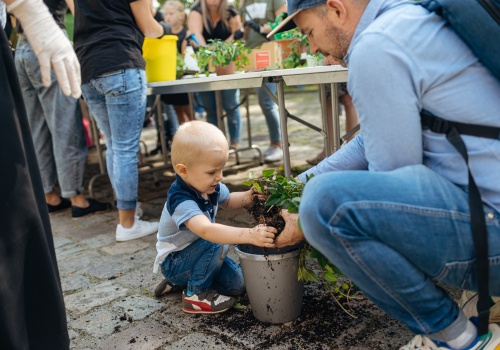 Zdjęcie, zbliżenie na dziecko kucające na ziemi, które próbuje umieścić zieloną roślinę w plastikowym pojemniku. Pomaga mu mężczyzna kucający tuż obok chłopca. W tle znajdują się inni uczestnicy warsztatów.