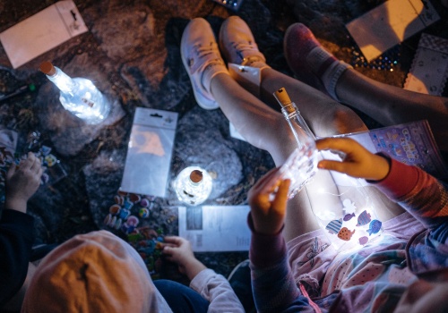 Zdjęcie, zbliżenie na kilka świecących latarenek. Latarenki są zbudowane z butelek i świecących się lampek. W kadr wkradają się nogi i ręce dzieci, które siedzą na ziemi.