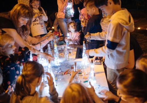 Zdjęcie, w kadrze znajdują się licznie zebrani uczestnicy festiwalu wokół stołu warsztatowego. Zapadł zmrok. Dzieci wraz z dorosłymi tworzą morskie, świecące się butelki.