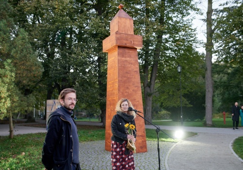 Fotografia przedstawiająca Pawła Błęckiego oraz Kingę Jarocką na otwarciu wydarzenia „PRZYSTAŃ. PAWEŁ BŁĘCKI”. Za postaciami znajduje się schematycznie wykonany obiekt z drewna, przypominający latarnię morską. 