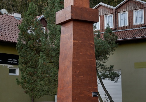Fotografia przedstawiająca schematycznie wykonany obiekt z drewna, przypominający latarnię morską, autorstwa Pawła Błęckiego. Za obiektem znajdują się niskie budynki oraz karłowate drzewa. Fotografia wykonana podczas wydarzenia „PRZYSTAŃ. PAWEŁ BŁĘCKI”.