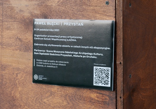 Zbliżenie na tabliczkę z informacjami o obiekcie autorstwa Pawła Błęckiego „PRZYSTAŃ”. W prawym dolnym rogu znajduje się kod QR.