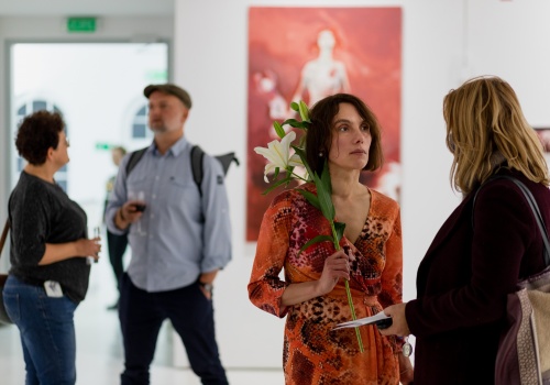 Zdjęcie. Kadr na artystkę – Annę Orbaczewską, trzymającą kwiat w dłoni. Artystka zwrócona jest w stronę kobiety z którą rozmawia. W tle ukazują się się kolejne postacie oraz obraz Anny Orbaczewskiej, wyeksponowany wewnątrz przestrzeni wystawienniczej CSW Łaźnia.