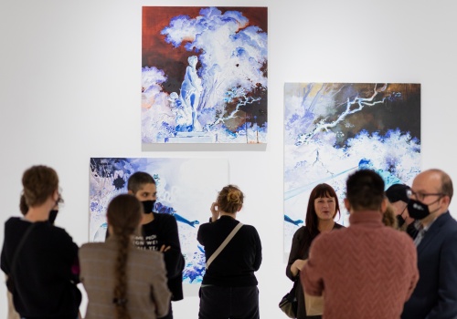 Fotografia przedstawia grupę ludzi na wystawie Anny Orbaczewskiej „Pułapki i Przynęty”. W tle ukazują się obrazy artystki, kolorystycznie przypominające negatyw.