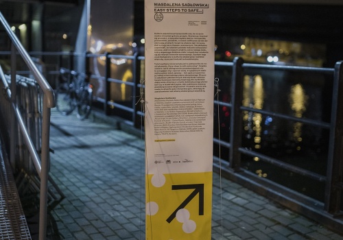 Zbliżenie na infografikę wydarzenia „Magdalena Sadłowska – Easy Steps to Safe…”, w formie standu. Po lewej stronie znajduje się fragment rampy prowadzącej do wejścia galerii LKW. Po prawej stronie, za barierką uchwycono rzekę Motławę. 