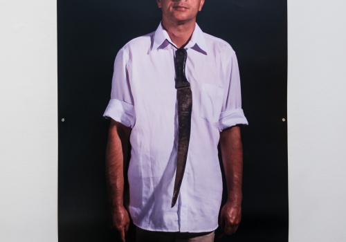 Fotografia Rosemberga Sandovala, wyeksponowana na sali wystawienniczej. Artysta stoi wyprostowany, ubrany w jasną koszulę. Pod kołnierzykiem postaci zawieszony jest duży, zardzewiały nóż, imitujący krawat. Czarne tło.