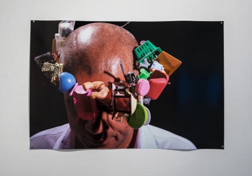 Fotografia z wizerunkiem artysty, który na głowie ma wianek skonstruowany z drobnych zabawek dla dzieci. Artysta ma pochyloną głowę. Czarne tło.