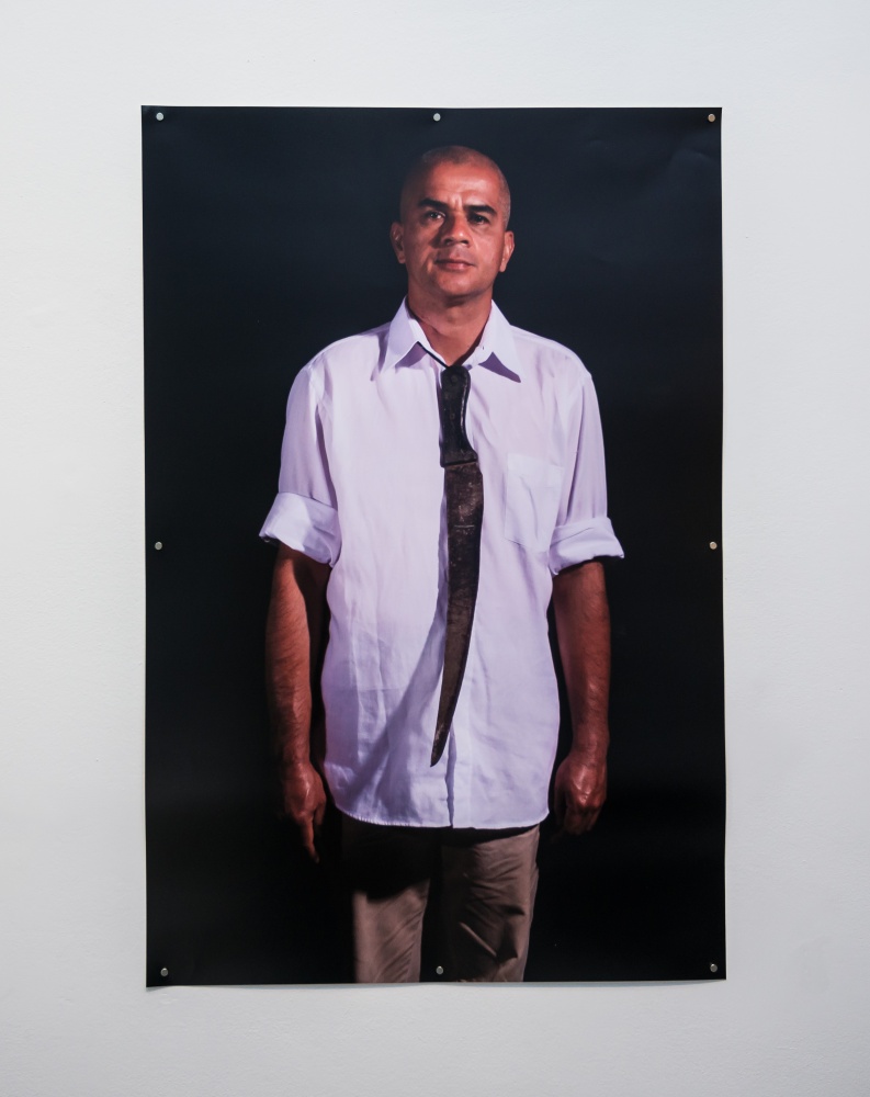 Fotografia Rosemberga Sandovala, wyeksponowana na sali wystawienniczej. Artysta stoi wyprostowany, ubrany w jasną koszulę. Pod kołnierzykiem postaci zawieszony jest duży, zardzewiały nóż, imitujący krawat. Czarne tło.
