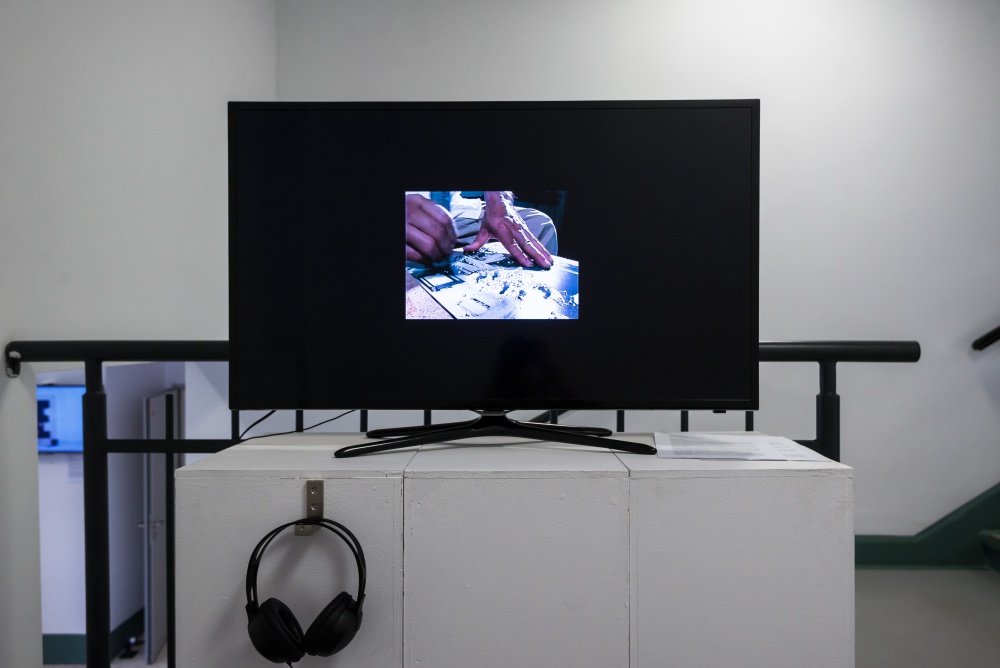 Kadr na białe kubiki z telewizorem, na którym wyświetlana jest praca audiowizualna autorstwa Rosemberga Sandovala. Na ekranie znajduje się fragment postaci bez widocznej twarzy.