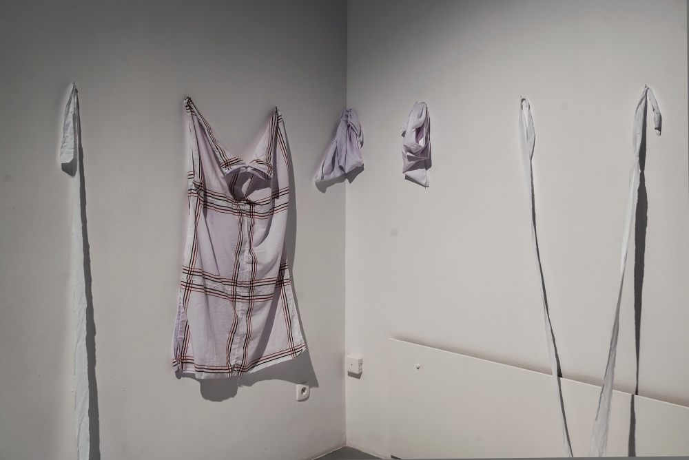 Kadr na instalację artystyczną, będącą pozostałością po wernisażowym performansie Rosemberga Sandovala. Jasne pasy tkanin przymocowane do ścian, swobodnie opadających na podłogę oraz zawieszona na ścianie prosta, jasna sukienka.