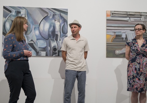 3.	Trzy osoby stojące przed białą ścianą w sali wystawienniczej. Na ścianie rozwieszone obrazy  przedstawiające fragmenty broni palnej.