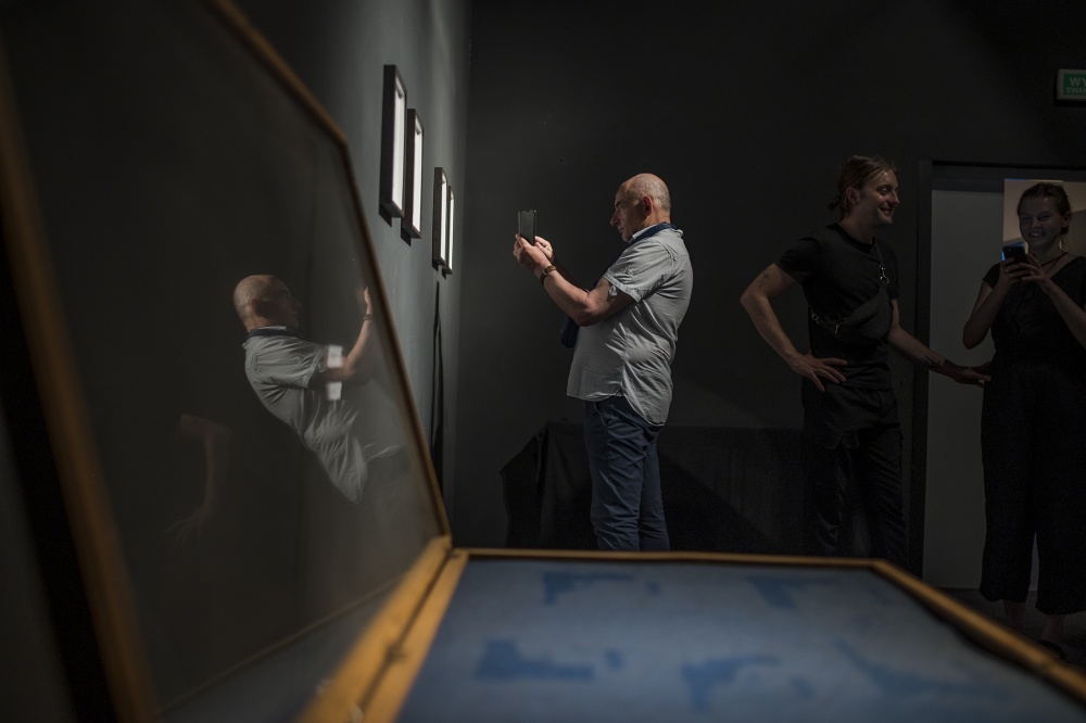 1.	Trzy osoby w ciemnej Sali wystawienniczej. Jedna z osób wykonuje fotografię jednej z prac za pomocą smartfona.