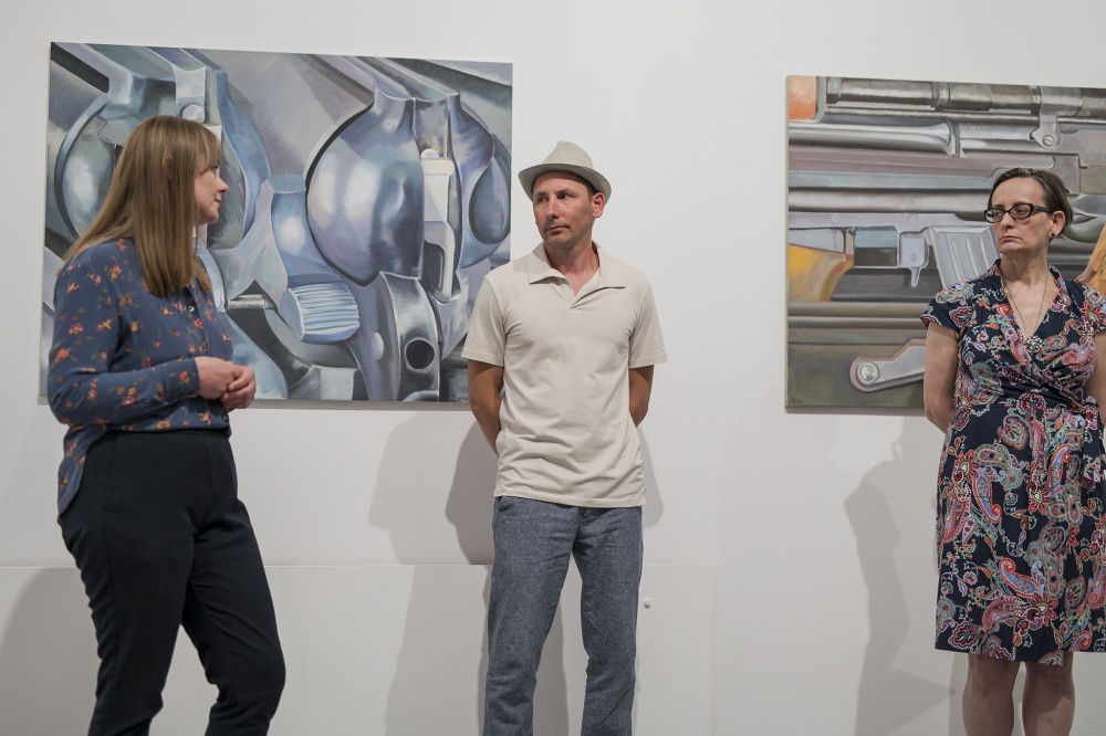 3.	Trzy osoby stojące przed białą ścianą w sali wystawienniczej. Na ścianie rozwieszone obrazy  przedstawiające fragmenty broni palnej.