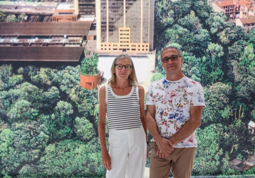 1.	Mężczyzna i kobieta w wieku średnim stojący na tle fototapety przedstawiającej panoramę miejską, częściowo zalesioną.