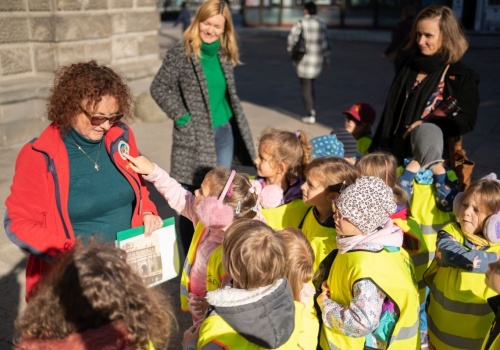 Grupa dzieci w odblaskowych kamizelkach, wraz z przewodniczką wycieczki przy Bramie Wyżynnej. Jedno z dzieci wskazuje na emblemat z herbem Gdańska, naszyty na bluzę przewodniczki.