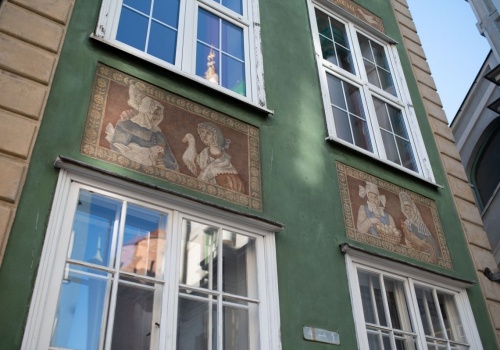 Zbliżenie na fasadę zielonej kamienicy z dużymi oknami oraz muralami postaci w starodawnych kostiumach
