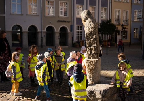 Kadr na grupę dzieci w odblaskowych kamizelkach, zebranych wokół kamiennej rzeźby przed Bazyliką Mariacką.