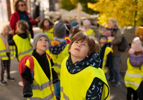 Zbliżenie na zamyślone twarze dzieci w odblaskowych kamizelkach, w trakcie trwania miejskiego spaceru.