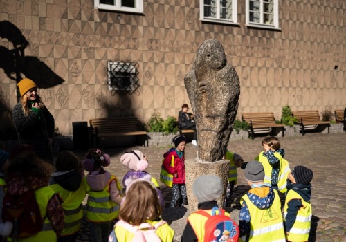 Kadr na abstrakcyjną rzeźbę, znajdującą się przed Bazyliką Mariacką. Wokół obiektu zebrane są dzieci w odblaskowych kamizelkach, biorące udział w spacerze miejskim.