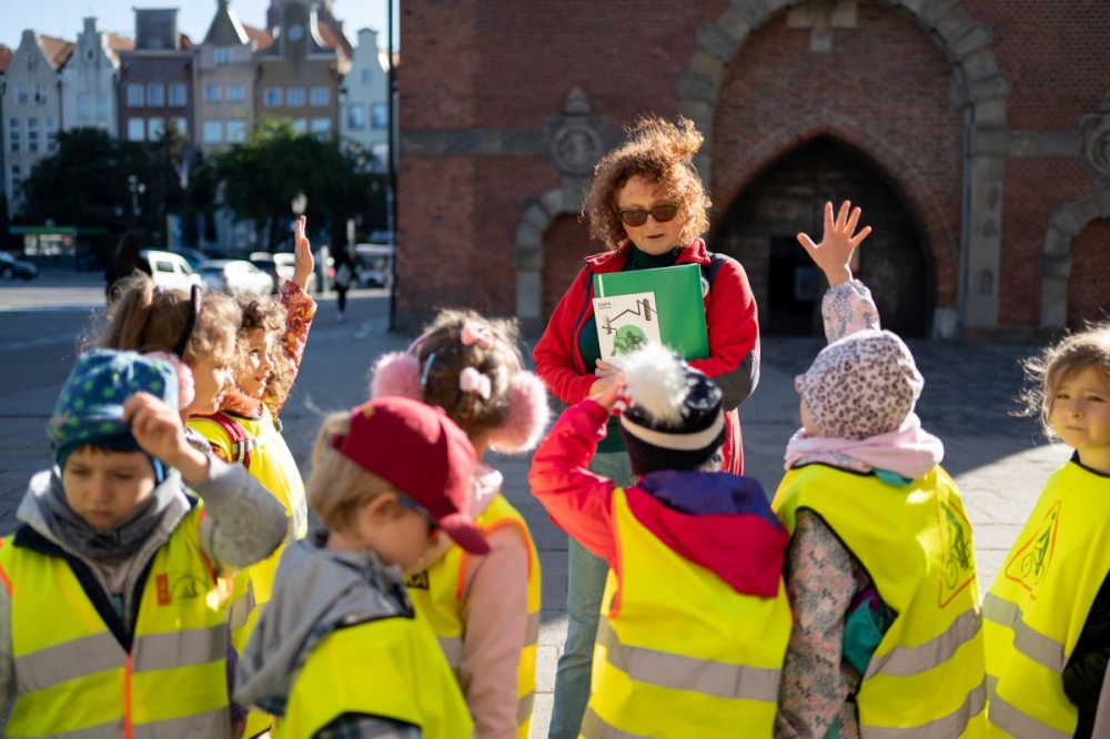 Grupa dzieci wraz z przewodniczką wycieczki na tle Katowni. Część z dzieci zgłasza się do odpowiedzi.