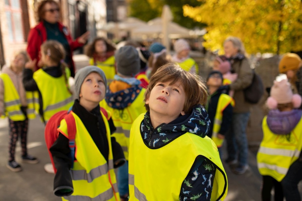 Zbliżenie na zamyślone twarze dzieci w odblaskowych kamizelkach, w trakcie trwania miejskiego spaceru.