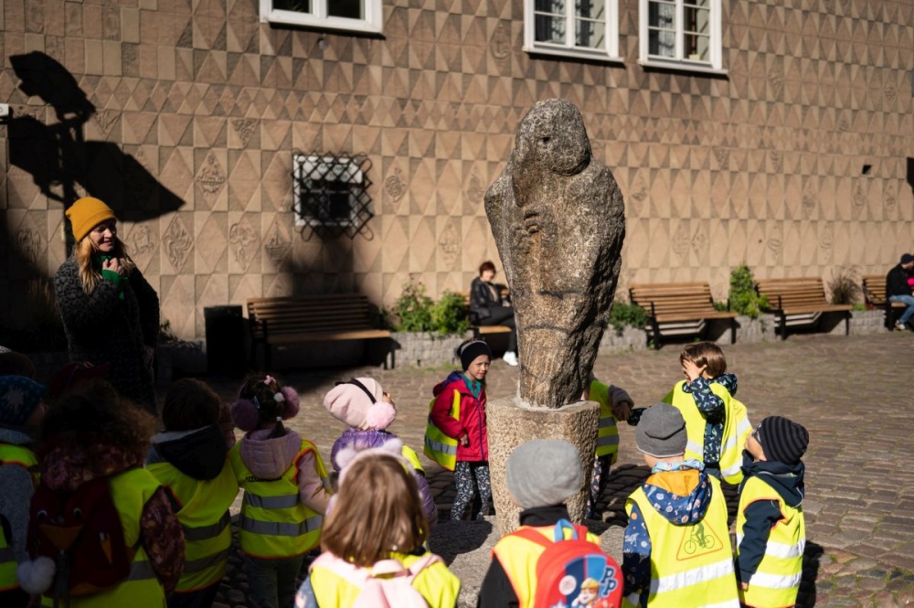 Kadr na abstrakcyjną rzeźbę, znajdującą się przed Bazyliką Mariacką. Wokół obiektu zebrane są dzieci w odblaskowych kamizelkach, biorące udział w spacerze miejskim.