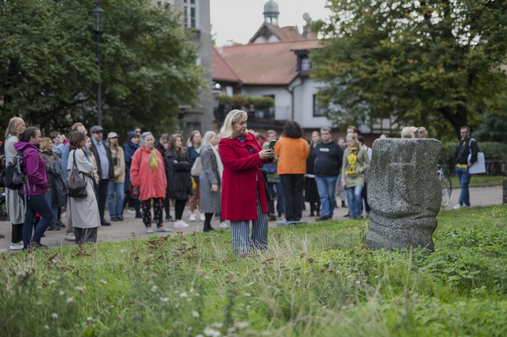 Liczna grupa ludzi przy abstrakcyjnej kamiennej rzeźbie w przestrzeni publicznej. Kobieta ubrana w czerwony płaszcz stoi najbliżej obiektu i fotografuje go za pomocą smartfona.