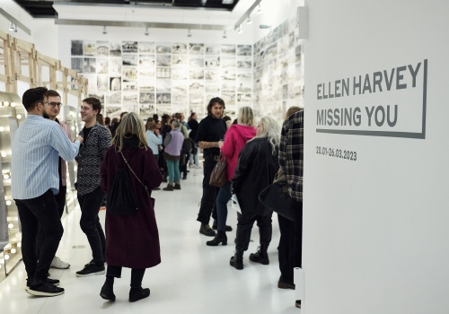 Zdjęcie. Publiczność  w sali wystawienniczej, w tle prace artystki Ellen Harvey. Z prawej strony widoczna plansza z tytułem wystawy „Ellen Harvey – Missing you”. 