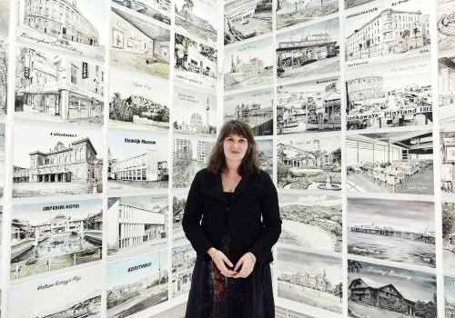 Zdjęcie. Portret artystki Ellen Harvey na tle pracy „Zawiedziony turysta” składającej się z obrazów rozwieszonych na całej powierzchni dwóch ścian.