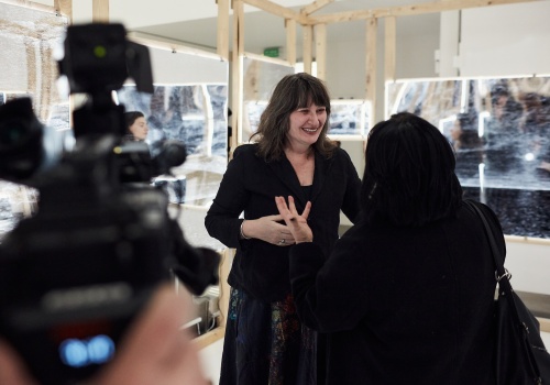 Zdjęcie. Artystka Ellen Harvey udzielająca wywiadu telewizyjnego wewnątrz instalacji „Arkadia”. Na pierwszym planie widoczna kamera telewizyjna.