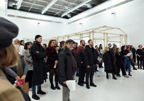 Zdjęcie. Publiczność zgromadzona w sali wystawienniczej w trakcie wernisażu. W tle widoczna drewniana konstrukcja instalacji artystycznej „Arkadia”.
