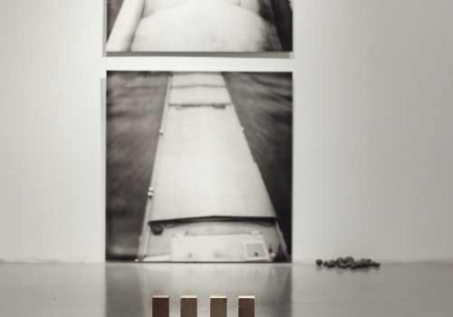 Zdjęcie. Perspektywa sali wystawienniczej. Na ścianie dwie fotografie w kwadratowym formacie, fotografie wiszące jedna nad drugą, dolna zaraz przy podłodze. Po prawej od fotografii instalacja artystyczna z małych szarych kamieni nieregularnie rozłożonych. Prace autorstwa Anny Kędziory.