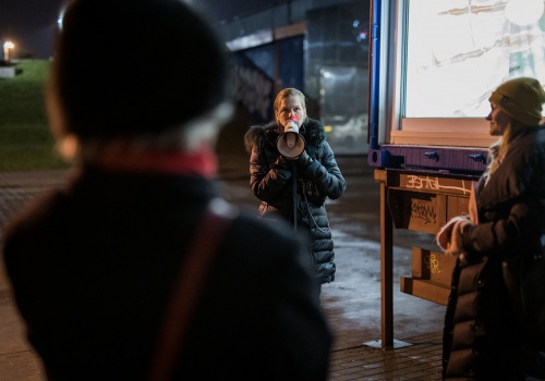 Fotografia pozioma wykonana nocą. Po środku artystka Marianna Serocka przemawiająca do megafonu. Po prawej stojąca bokiem Anna Szynwelska, kuratorka wystawy. Za kobietami widać rozświetlone wnętrze TIR-a, a na dalszym planie zarys wiaduktu.
