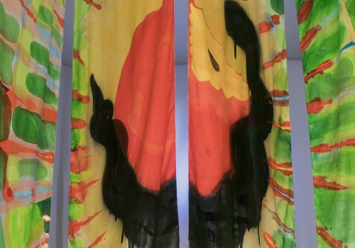 Fotografia pionowa przedstawiająca pracę „Wonsz” wewnątrz kontenera LKW Gallery. Na zdjęciu widać cztery pionowe, prostokątne panele z tkaniny, pomalowane na jasne, żywe kolory. W centralnej części obrazu czarny wąż oplatający czerwone jabłko, a dookoła pomarańczowe i zielone promienie na białym tle. 
