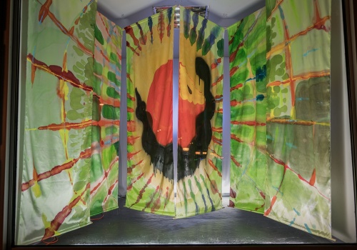 Fotografia pozioma przedstawiająca pracę „Wonsz” wewnątrz kontenera LKW Gallery. Na zdjęciu widać sześć pionowych paneli z tkaniny, pomalowanych na jasne, żywe kolory. W centralnej części obrazu czarny wąż oplatający czerwone jabłko, a dookoła pomarańczowe i zielone kształty przypominające promienie na jasnym tle. 