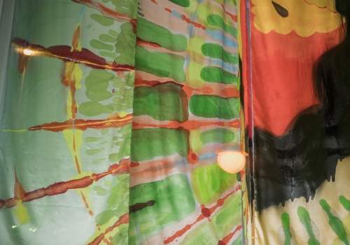 -- Fotografia pionowa przedstawiająca lewy fragment pracy „Wonsz” wewnątrz kontenera LKW Gallery. Na zdjęciu widać trzy pionowe panele z tkaniny, pomalowane na jasne, żywe kolory (czerwony, zielony, żółty) w psychodeliczne wzory przypominające promienie lub orbity planet.