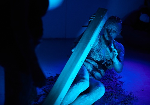 Zdjęcie. W sali wystawienniczej oświetlonej na niebiesko, ubrana w czarny kostium i wysmarowana gliną performerka Monika Wińczyk, kładzie się na podłodze, przykrywając kłodą drewna. 