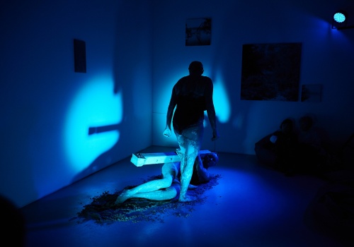 Zdjęcie. W sali wystawienniczej oświetlonej na niebiesko Monika Wińczyk i Magdalena Mellin wykonują performans. Artystki są ubrane w czarne kostiumy i wysmarowane gliną. W tle widać osoby z widowni, a na ścianach fotografie z wystawy.