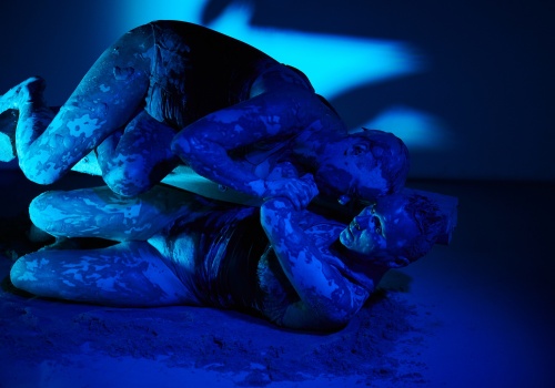 Zdjęcie. Zbliżenie na leżące na sobie performerki Monikę Wińczyk i Magdalenę Mellin, wykonujące performans w oświetlonej na niebiesko sali wystawienniczej. Artystki są ubrane w czarne kostiumy i wysmarowane gliną.